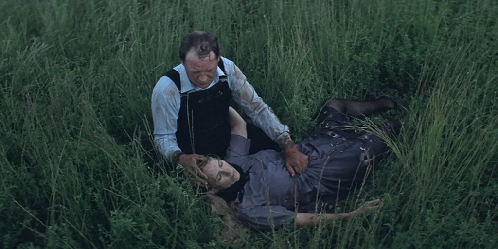 A man crouches on the ground holding a woman in a grass field in a still from Silent Light