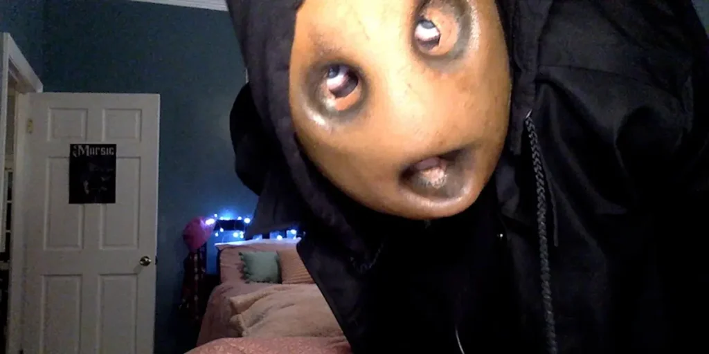 A person wears a disquieting mask with big eyes and an open mouth and a cape in a still from the movie Strange Harvest