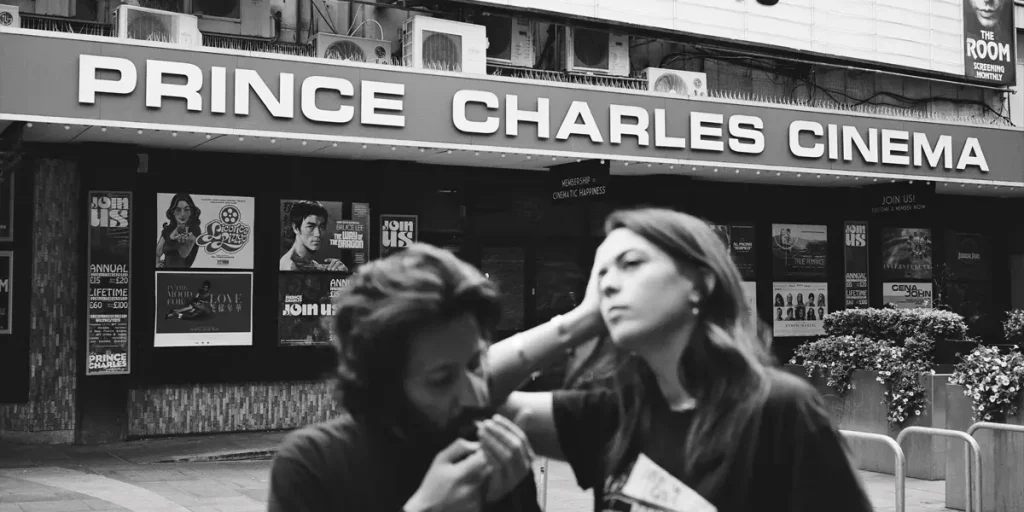 Two people stand in front of the Prince Charles Cinema in a the poster of the film The Regulars (2025)