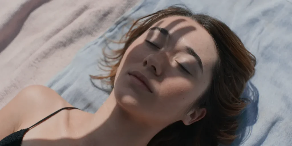 A young woman lies on a towel with the shadow of a hand on her face blocking the sun in a still from the movie Forastera