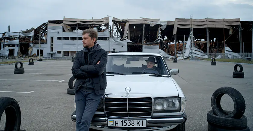 A man stands next to a Mercedes car with another man sitting inside in a still from To The Victory!