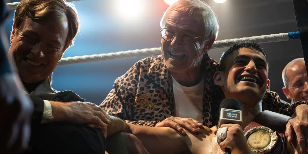 A boxer is in the ring, holding a microphone with happy people around him, in a still from the movie Giant