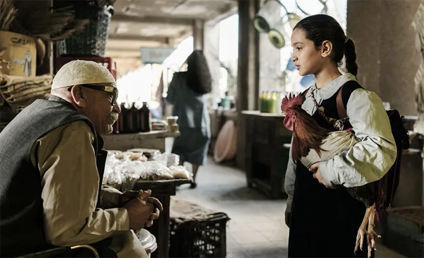 A girl holds a chicken while speaking to a man in a still from The President's Cake
