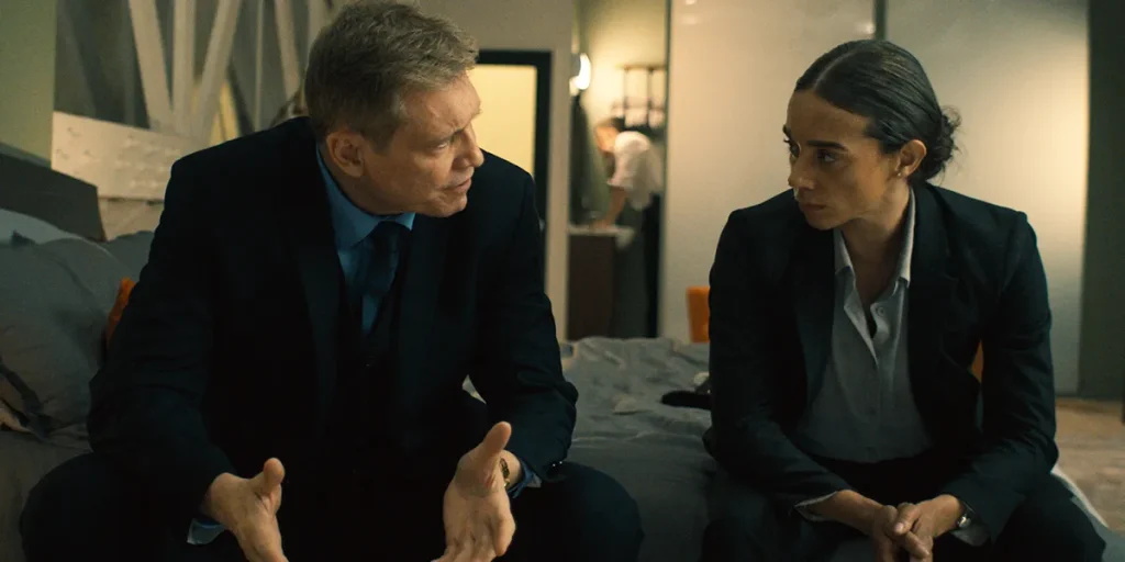 A man and a woman sit on a bed talking to each other in a still from the 2025 movie "Safe House"