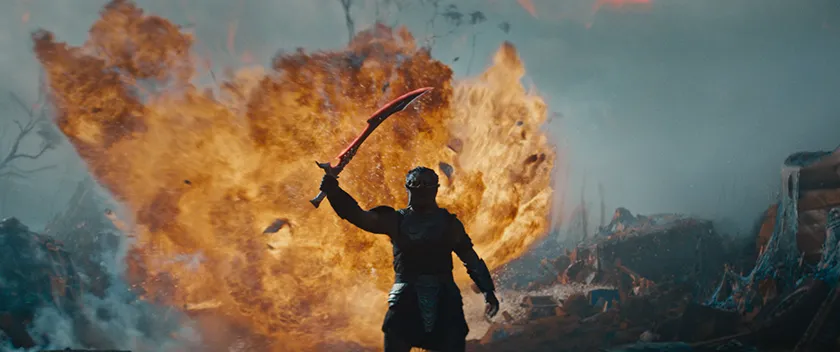 A creature wields a sword standing in a wasteland with fire behind them in a still from the movie Predator: Badlands