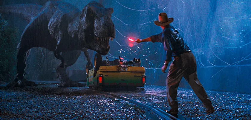 A dinosaur towers over an upside down car with a man in front of him in a still from Jurassic Park (1993)