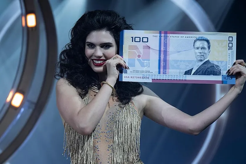 A woman holds an Arnold Schwarzenegger 100$ note in a still from the new Running Man movie (2025), which is better than the original according to Loud And Clear Reviews