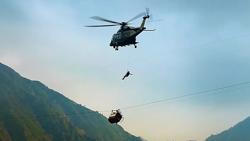 A man hangs from a helicopter towering above a cable car in a still from Hanging by a Wire (2026)