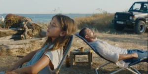 A girl and a man, father and daughter, lean on two deckchairs facing opposite ways in a still from the 2026 movie Hold Onto Me (Κράτα Με)