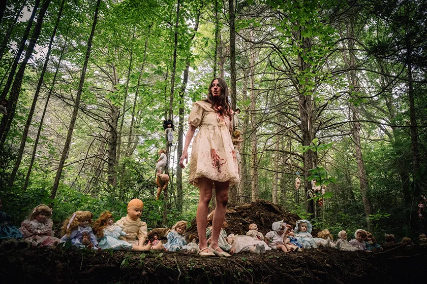 Fabianne Therese stands in a forest wearing a a blood-smeared white dress, with a row of ceramic dolls on the ground and others on the trees in Rod Blackhurst’s Dolly