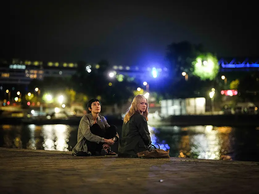 Two women sit by the river, the blonde one looking at the camera and the dark-haired one looking up, in a still from Ryūsuke Hamaguchi's All of a Sudden (Soudain)
