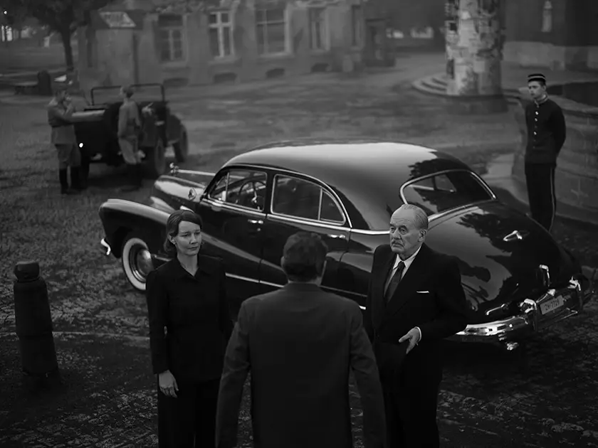 Hanns Zischler and Sandra Hüller stand in front of a car talking with someone in Paweł Pawlikowski's film Fatherland