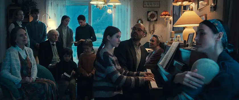 Sebastian Stan and Renate Reinsve sit, with one of their children playing piano and the rest of their family around them, in a still from the movie Fjord