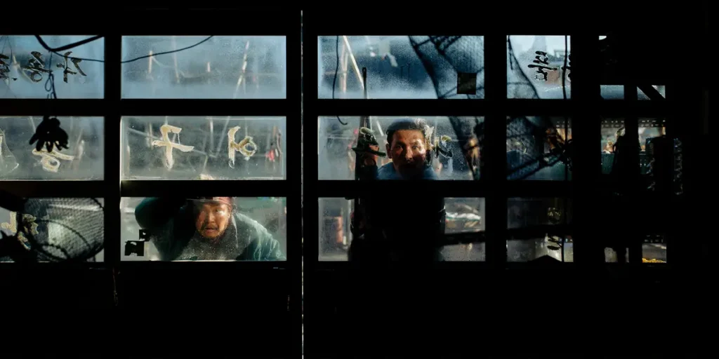 Two people stand behind a glass door that we see from inside, in the darkness, in Hope (호프), from director Na Hong-Jin