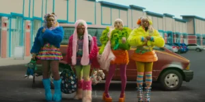 Four women stand in front of a car wearing eccentric clothes in a still from the movie I Love Boosters