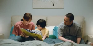 Mother and father sit on a bed with their child in the middle, reading a story from a yellow book, in Sheep in the Box (箱の中の羊 - Hako No Naka No Hitsuji)