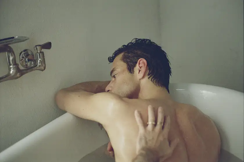 A hand touches Rami Malek's back while he's in a bathtub in The Man I Love