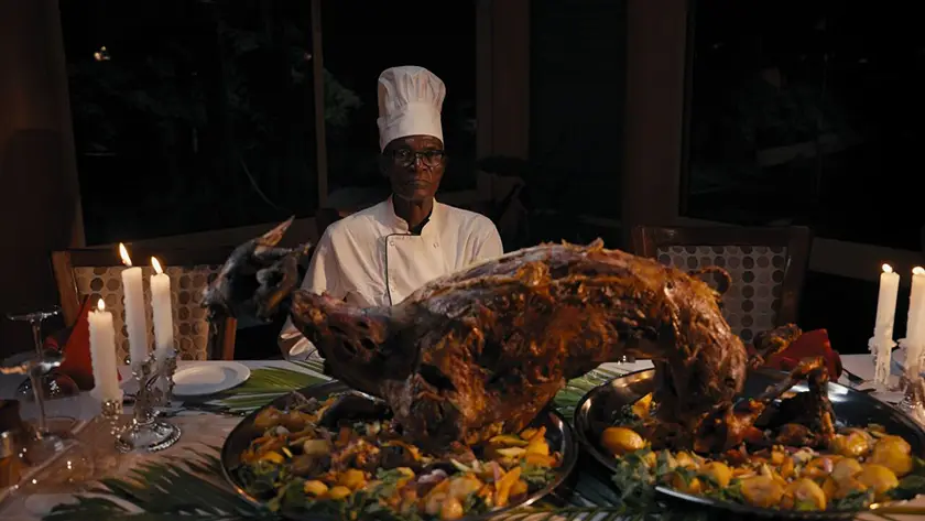 A chef sits by a table with a lot of food in front of him in a still from the documentary How to Feed a Dictator