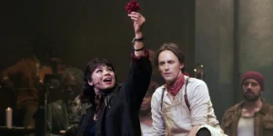 Eurydice (Eva Noblezada) holds up a red flower with Orpheus (Reeve Carney) sitting next to her, looking at her, in a still from the 2026 movie "Hadestown: The Musical"