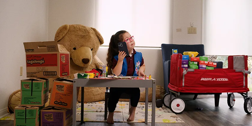 5-year-old Ara looks serious on the phone, sitting at a tiny desk, with boxes of cookies next to her and a giant teddy bear behind her in a still from the movie Cookie Queens