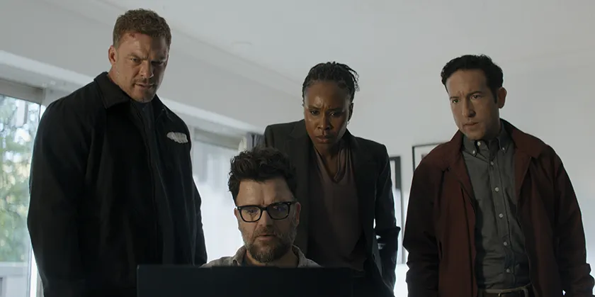 Four people look at a computer screen, one of them sitting and typing, in a still from Reacher Season 4, which this article explores everything we know about