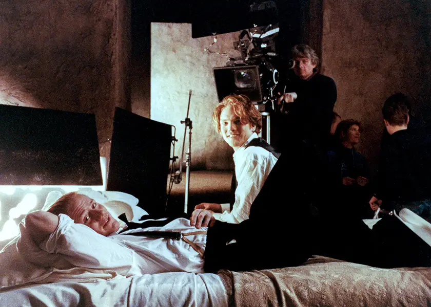 A young man sits on a bed next to another young man who's lying down on it, with another man filming them, in a still from the movie Queer Edward II
