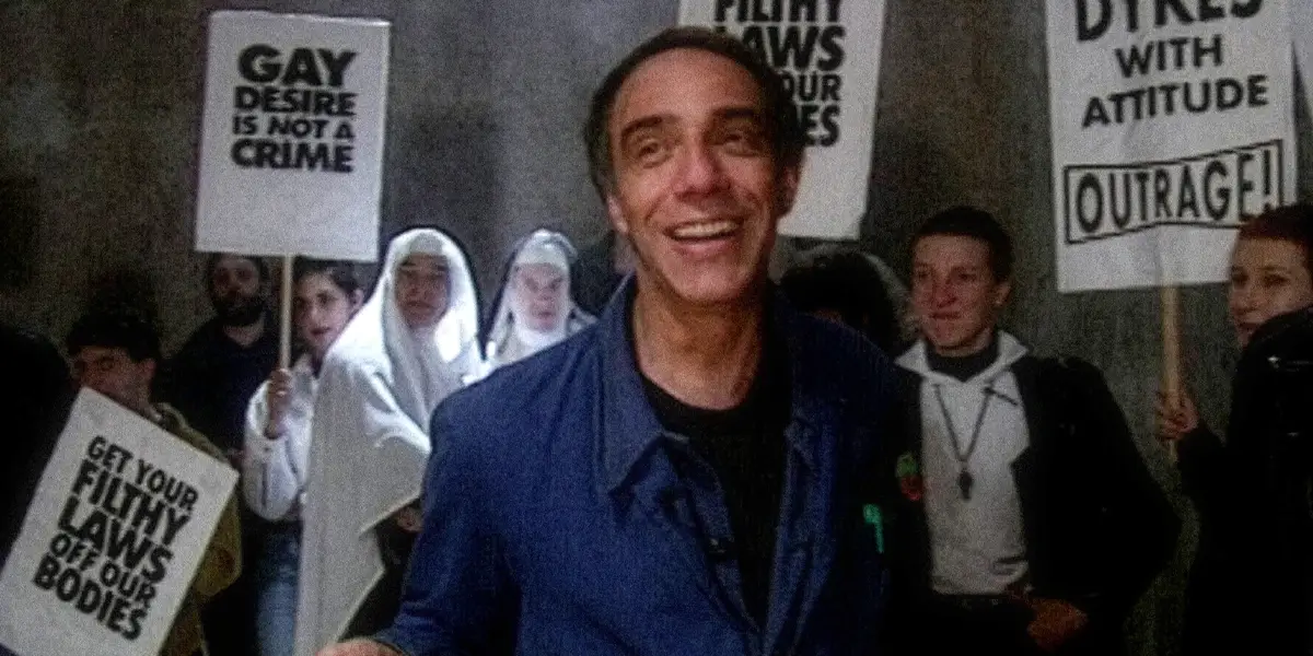 A man in a blue shirt stands in front of a crowd of people holding signs in a still from Queer Edward II, one of the 30 movies to watch at the 2026 BFI London Film Festival according to Loud and Clear Reviews