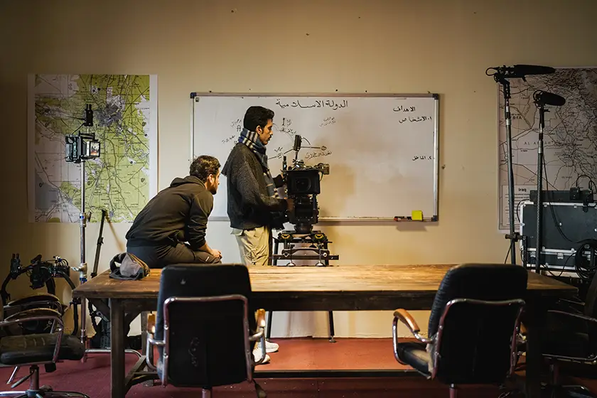 Two film crew members preparing a professional camera inside a production studio, surrounded by filming equipment.